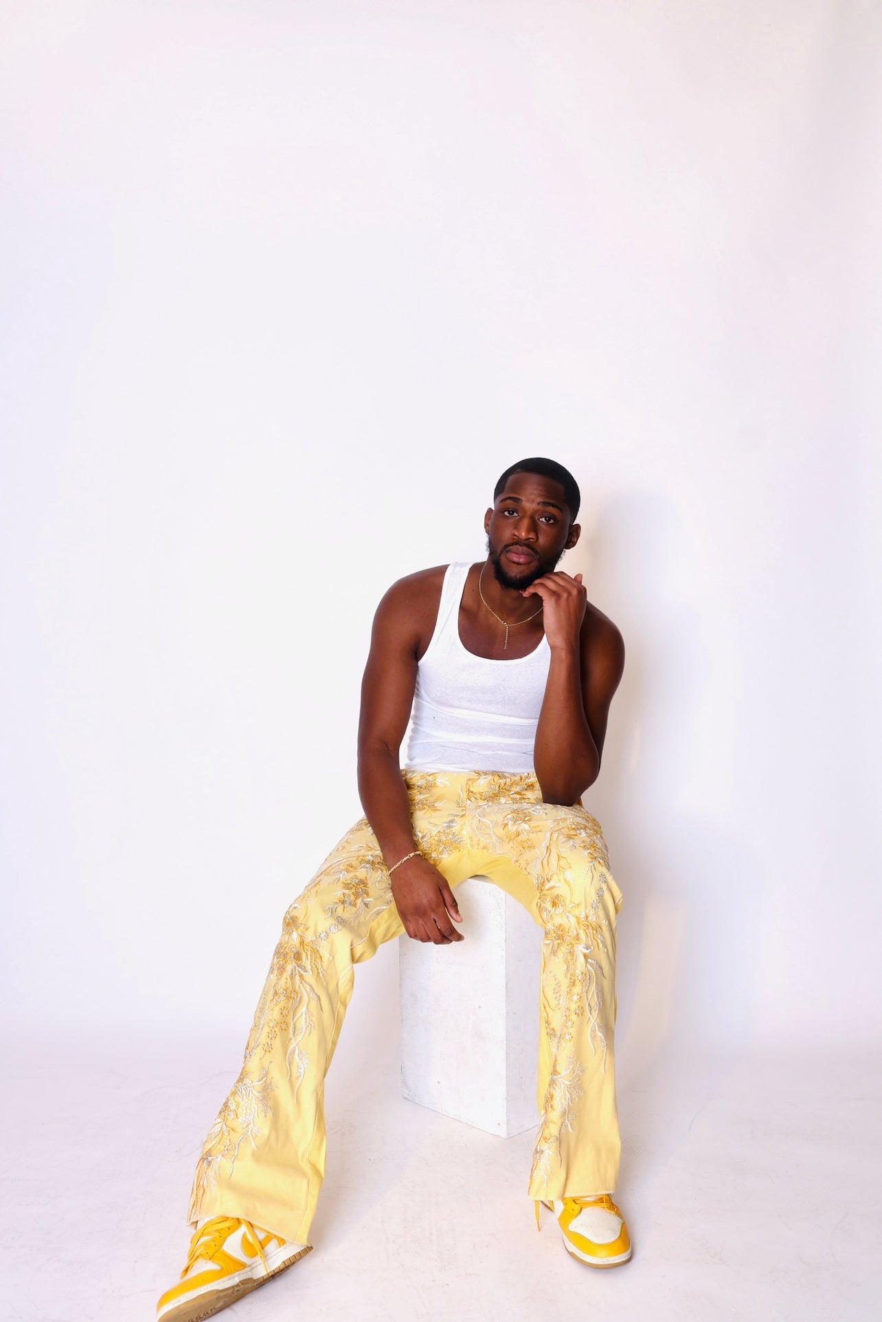 Person wearing a white tank top and yellow pants sitting on a white stool against a white background